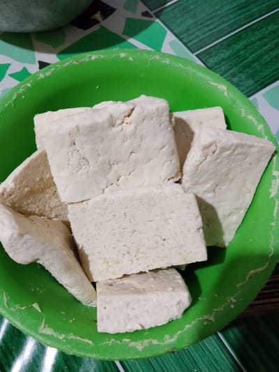 A close-up, overhead shot shows pieces of white tofu piled in a bright green plastic bowl. The tofu is cut into irregular, rectangular shapes, some appearing slightly crumbly. The bowl is placed on a surface with a patterned green and white checkered design. The lighting is dim, casting shadows on the tofu. The location is Merbopuntong, Indonesia.