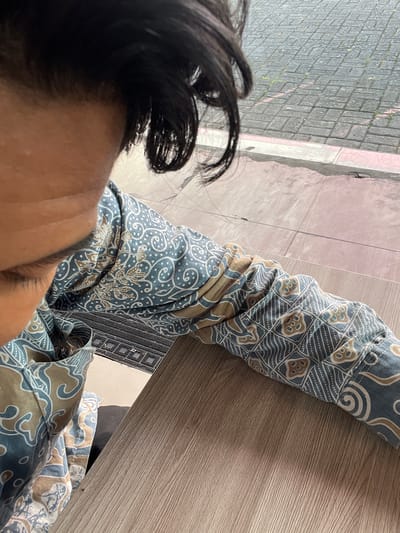 This image captures a close-up, overhead view of a person's head and arm, draped in a batik-patterned garment. The fabric features intricate designs in blue, beige, and white, with swirling motifs and geometric shapes. The person's dark hair is visible on the left side of the frame, styled loosely and falling forward. The arm, also covered in the batik fabric, rests on a wooden surface, likely a table, which occupies the right side of the image. The wood grain is clearly visible, suggesting an indoor setting or a covered outdoor area.

In the background, to the left of the arm and below the person's head, a textured surface of grey rectangular tiles is visible. Faint pink lines are present on the tiles, possibly indicating grout lines or a subtle pattern. A lighter colored strip, possibly a curb or border, runs alongside the tiled area. A glimpse of a darker textured mat with a diamond pattern can be seen beneath the arm and the wooden surface.

The lighting suggests it is daytime, with diffused light creating soft shadows. The overall impression is one of casual relaxation. The location context of Tampingan, Indonesia, combined with the batik garment, points towards a cultural setting. No text is visible in the image.