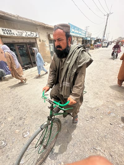 In Bostan, Pakistan, a man with a beard and a skullcap rides a green bicycle on a dusty, paved street. He wears a brown outfit with a large, draped scarf. Several people are visible in the background, some walking and others on motorcycles. Shops line the street, including one labeled "NOOR X-RAYS. Kucha," and another with "AL NISAR PHARMACY" written in blue lettering. Power lines crisscross overhead. The sky is bright and overcast.