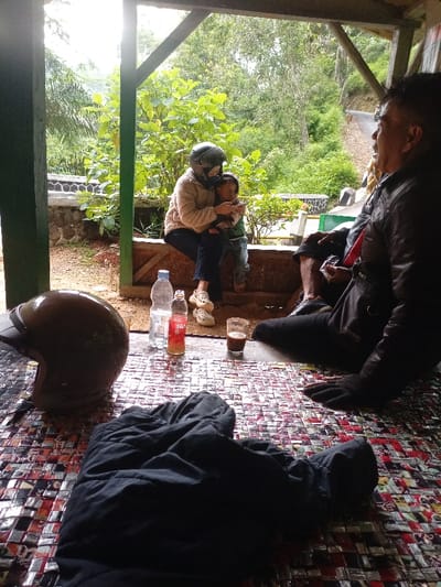 The image shows a group of three people sitting on a tiled floor outside a structure in Cisewu, Indonesia. The location is determined by the coordinates provided, which correspond to an area in Cisewu. One person, a woman wearing a helmet and beige jacket, is sitting and holding a young child on her lap. The child is looking at a screen, possibly a phone, and holding a small object in their hand. The woman is wearing black pants and white shoes. Another person, an older man, is sitting to the right of the woman. The man is wearing a black leather jacket, black pants, and a white shirt. He is looking in the direction of the woman, and he has a brown bag strapped across his body. The man has a black glove on his left hand, which is resting on the tiled floor. The three people are all looking at each other. There are two glass bottles and a glass cup with a brown liquid on the tiled floor in front of them. The scene is a casual outdoor setting.  The floor is covered in a tile pattern, and there are two open wooden structures behind the individuals, likely the remains of a small house or a porch.  The floor is also covered with discarded cigarettes, cigarette wrappers, and a black motorcycle helmet.