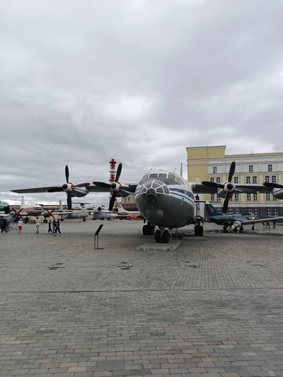 An overcast day at an outdoor museum in Verkhnyaya Pyshma, Russia, features several aircraft on display. The primary subject is a large, four-engine turboprop aircraft with a grey and white livery and visible lettering in Cyrillic. It is parked on a paved plaza. To its right, a smaller, dark blue jet aircraft with a red star insignia is visible. In the background, a multi-story yellow building with white-trimmed windows stands behind the aircraft. Other aircraft, including a red and white propeller plane, are positioned further back. A few individuals, including a group walking together, are scattered across the plaza. The ground is paved with rectangular stone tiles.