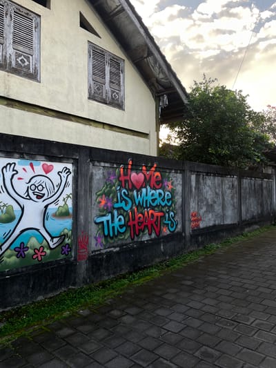 The image shows a section of a wall with graffiti in the city of Kediri, Indonesia. The wall is concrete and runs alongside a paved walkway. The graffiti on the left depicts a white stick figure with its arms raised in excitement, set against a backdrop of islands, palm trees, and a blue sky with clouds. Red hearts are above the figure's head. To the right of this, graffiti spells out "Love is where The Heart is" in colorful, blocky letters with floral designs. The wall itself is textured and shows signs of age and weathering. Behind the wall, a building with weathered wooden shutters on two windows is visible, and a lush green tree grows near the corner of the building. The sky above is partly cloudy, with sunlight filtering through. The paved walkway in the foreground appears wet, suggesting recent rain. The overall scene suggests an outdoor urban environment with vibrant street art.