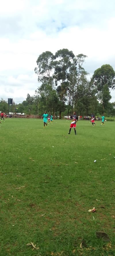 The image shows a soccer field in Lubao, Kenya, located at latitude 0.384, longitude 34.801. The field is covered in lush green grass, with a few scattered leaves. There are nine people on the field, all wearing soccer jerseys. The players are engaged in a game of soccer. In the background, there are trees and a cloudy sky. The players are wearing various colored jerseys, and the field is surrounded by a fence. The environment is predominantly green, with a few scattered patches of brown dirt. One player wearing a black and red jersey stands with his legs apart and his hands on his hips. Another player is in mid-stride while running toward the left side of the frame.