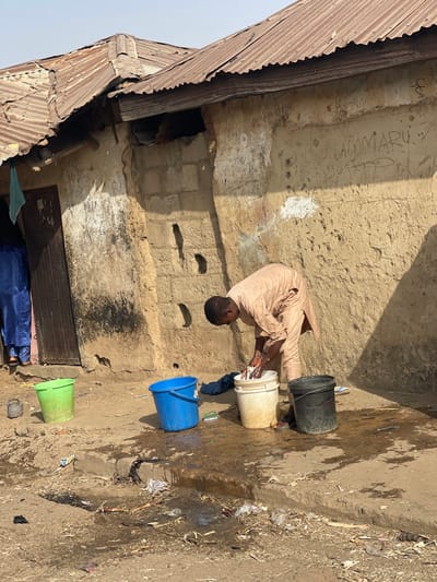 The scene depicts an outdoor activity in Unguwar Gabas, Nigeria, occurring under clear daylight. A male individual, dressed in a light-colored tan or beige tunic, is bent over, actively engaged in hand-washing items within a white plastic bucket. His hands are submerged in the water inside the bucket. Surrounding him on the uneven, dirt ground are two other plastic buckets: one blue, partially filled with liquid, and one black. A green plastic bucket and a small dark pot are positioned further left, near a wooden doorway. A blue fabric item and a small scrub brush are visible on the ground near the blue bucket. Water spillage from the washing activity is evident on the dirt and concrete ground, mixing with scattered debris, including plastic bags and wrappers.
The background consists of a weathered building with walls constructed from mud bricks and possibly some concrete blocks, exhibiting discoloration and erosion. The word "UMMARU" is visible as graffiti etched into the upper wall. The building is topped with a rusty, corrugated metal roof. A second individual, wearing a blue garment, is partially visible standing inside the doorway to the left, observing the scene.