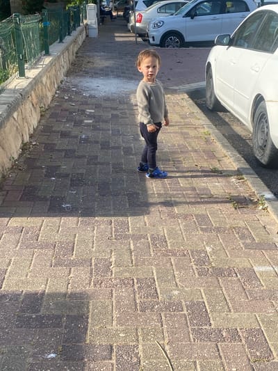 A child stands on a brick sidewalk in Kiryat Bialik, Israel. The child is wearing a gray sweater, dark pants, and blue sneakers. To the left are parked cars and a green fence. The sidewalk is composed of brown and gray bricks. The scene is lit with sunlight, casting shadows.