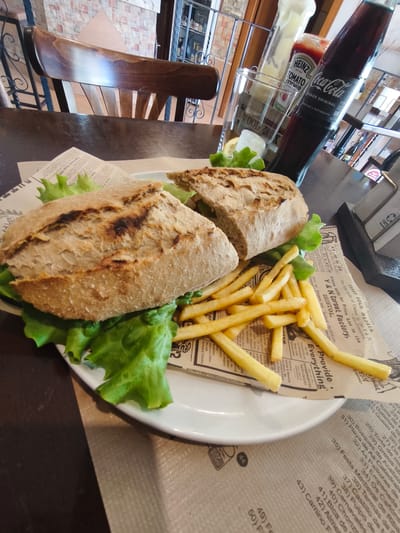 The image presents a close-up view of a casual meal served in an indoor establishment, likely a restaurant or cafe in Pontevedra, Spain. The main subject is a large, rustic-style sandwich, cut in half, featuring a hearty crusty bread and ample green lettuce. It is served on a white plate alongside a generous portion of golden-brown french fries.
Accompanying the meal is a glass filled with ice and a slice of lemon, a bottle of Coca-Cola, a Heinz Tomato Ketchup bottle, and another white condiment bottle, likely mayonnaise. The Coca-Cola bottle prominently displays "SABOR ORIGINAL" (Original Flavor) and "Deliciosa y Refrescante" (Delicious and Refreshing) in Spanish.
The food is placed on a dark wooden table, partially covered by a lighter-colored mat. Under the plate, a decorative sheet of paper, resembling an old newspaper, serves as a liner. This paper features various English texts such as "Opportunity Book FREE," "THREE GOLD MEDALS," "Y & N Corset Factory, BRISTOL," and "Provide Everything."
In the background, a dark wooden chair with a slatted back is visible. Beyond that, a tiled wall with a geometric pattern and shelves stocked with bottles suggest a bar or dining area. On the right side of the image, a menu or list in Spanish is partially visible, featuring numbered items such as "Torre de," "Queso de," "Empanada," "Pulpo de," and specific Galician place names like "Playa de As Catedrais," "Bosques de Acebo," and "Pailozas," firmly establishing the regional location within Spain. No people are visible, and the time of day cannot be determined from the indoor setting. The scene portrays a ready-to-eat meal.