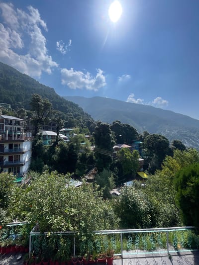 This image captures a panoramic view of a mountainous landscape in Bhagsu, India, under a clear, sunlit sky. The bright sun, positioned in the upper right, casts a strong glare. The sky is a vibrant blue with scattered white cumulus clouds.

In the foreground, a dark-tiled terrace is visible, featuring a green metal railing with decorative scroll patterns. Along the edge of the terrace, numerous red potted plants are arranged. Immediately beyond the railing, dense green foliage and large bushes obstruct a clear view of the lower ground.

The midground is dominated by lush, verdant mountains densely covered with trees. Several multi-story buildings are nestled into the mountainside. On the left, a prominent white building with multiple balconies featuring brown railings and green awnings is visible. Further into the scene, smaller houses with colored roofs (including teal and red) are interspersed among the trees. Utility lines stretch across the midground, suggesting infrastructure.

The background consists of successive layers of mountains, appearing lighter in shade due to atmospheric haze, extending into the distance under the clear sky. No people or overt actions are depicted in the scene.