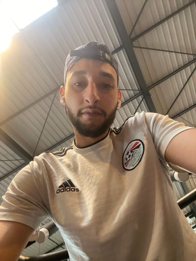 One male individual is depicted in a close-up, upward-angled self-portrait. He has short dark hair, a beard, and light-toned skin, and is looking directly into the camera. He is wearing a white short-sleeved t-shirt with a black Adidas logo on the right chest and a circular emblem on the left chest. The emblem contains a stylized soccer ball, text, and colors resembling the Egyptian flag, with the word "EGYPTIANYA" partially visible. Black stripes are present on the shoulders of the shirt. He is also wearing a dark-colored baseball cap reversed on his head and white wireless earbuds in both ears. The background consists of an indoor setting featuring a corrugated white ceiling with visible dark metal support beams and trusses. A bright light source is present in the upper left portion of the frame. The specific location within the city of Hœnheim, France, cannot be determined from the visual information provided.