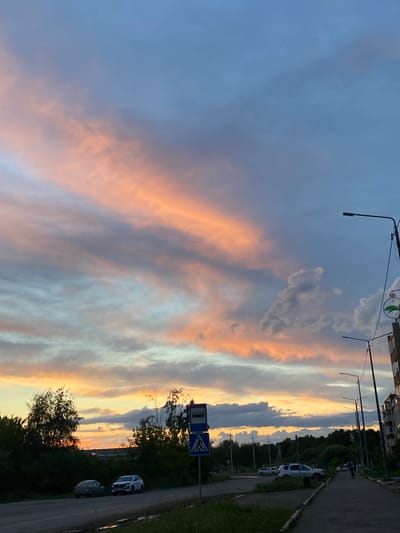 This vertically oriented photograph captures a sunset scene in Uchaly, Russia. The sky is filled with dramatic clouds, displaying hues of pale blue, soft orange, and light yellow, indicating the sun is low on the horizon. Streaks of warm color paint the sky, suggesting a picturesque sunset.
In the foreground and to the right, a paved road runs alongside a grassy verge and a line of trees. Several parked cars are visible along the roadside, including a white SUV and a darker colored car further down. There are also streetlights and what appear to be apartment buildings or other structures on the far left, hinting at a semi-urban or suburban environment.
A blue traffic sign with a white pictogram of a bus and a pedestrian crossing sign with a blue border and white background are prominently placed on the right side of the road. The overall atmosphere is serene and tranquil, typical of an evening setting during or after sunset. The lighting suggests it is late evening.