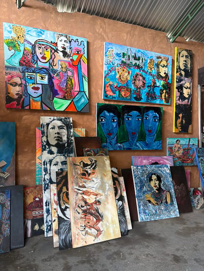 The image shows an art studio with many paintings displayed on a rough, terracotta-colored wall and leaning against it on the concrete floor. The paintings are varied in style and subject matter.
Prominently displayed on the wall are three large paintings. The leftmost one is a colorful, cubist-style collage of faces and figures. To its right is a vibrant blue painting with various figures, including what appear to be deities and people on bicycles, set against a backdrop of buildings. The third large painting on the wall is a vertical stack of three portraits, done in a graphic, black and white, and pink style.
In front of the wall, leaning on the floor, are numerous other paintings. These include a series of portraits, a painting of three blue-faced figures, a scene of a person in a boat, and several paintings with abstract designs and what appear to be animal motifs, such as a tiger's face and a group of horses in motion. Some paintings are stacked on top of each other, obscuring parts of the lower layers.
The environment appears to be an indoor space, possibly a workshop or gallery, with a corrugated metal roof visible at the top of the image. The lighting suggests it might be daytime, with natural light filtering in.
The location of this scene is identified as Tejakula, Indonesia, suggesting it is an artist's studio in that region.