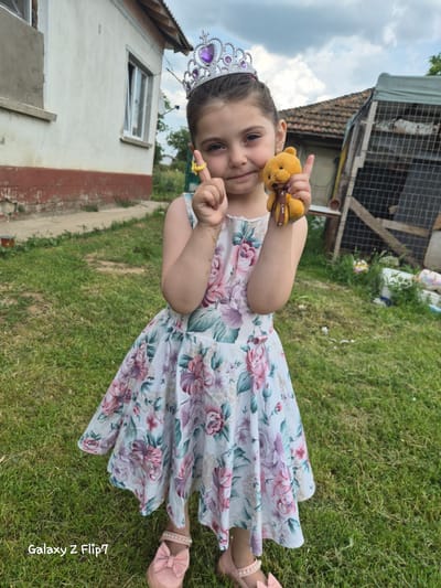 A young girl stands outdoors in Kalipetrovo, Bulgaria, looking directly at the camera with a slight smile. She is wearing a silver crown adorned with purple heart-shaped and circular jewels, a white floral sleeveless dress featuring pink, purple, and green patterns, and pink shoes with a bow and pearl-like embellishments. In her right hand, she holds a small yellow teddy bear with a brown ribbon inscribed "Love" around its neck. Her left index finger, adorned with a yellow ring, is pointed upwards. The immediate foreground consists of green grass. Behind her to the left, part of a white house with a red-brown base and a partially open white-framed window is visible. To the right and rear, a rustic structure with a brown tiled roof and a dark grid-like fence is present, suggestive of an enclosure. The sky above is overcast. A "Galaxy Z Flip7" watermark is located in the bottom left corner of the image.