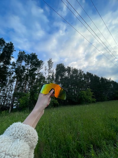 An outdoor, eye-level, wide shot depicts a hand holding a pair of iridescent, mirrored sunglasses up to the sky. The sunglasses are black-rimmed with a large, singular lens that reflects the colorful sky and surrounding trees. The hand, with its arm clad in a fluffy white jacket, is positioned in the lower-left quadrant of the frame. The background consists of a dense line of tall pine trees with dark green foliage, stretching across the middle of the image. In front of the trees, a field of tall, green grass, dotted with small white wildflowers, occupies the lower half of the frame. Above the trees, a cloudy, blue sky fills the upper portion of the image. Several power lines, running diagonally from the upper-right to the upper-left, are visible against the sky. The scene is set in a natural, rural environment, possibly a meadow or field adjacent to a forest, under a dynamic sky. The location is Podshivalovo, Russia.