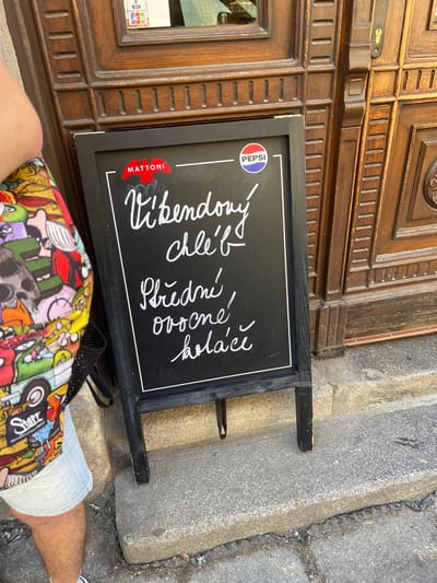 A chalkboard sign with white lettering stands in front of a wooden door. The sign advertises "Víkendový chléb" (Weekend bread), "Ovoce" (fruit), and "Koláče" (cakes). The Mattoni and Pepsi logos are visible on the sign. A person wearing a colorful graphic-print shirt and denim shorts stands to the left of the sign, partially obscuring it. The wooden door appears to be old and ornate, with carved details and a lock. The ground in front of the door is paved with stone. The scene is outdoors, likely on a street or sidewalk.