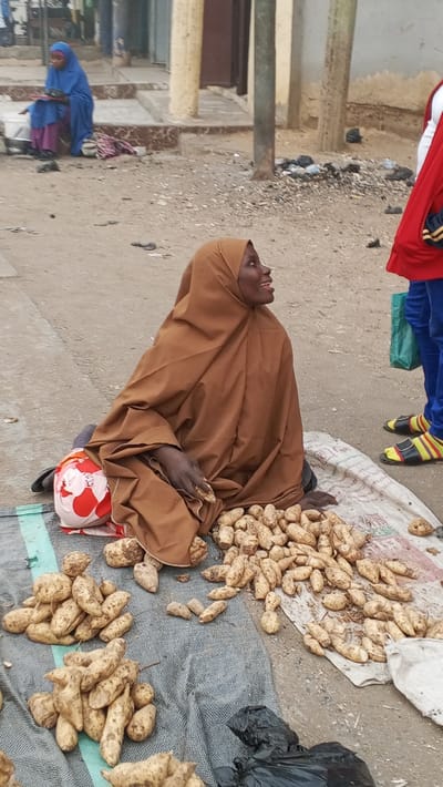 This image captures an outdoor street or market scene in Gawuna, Nigeria, centered around a female vendor.
The main subject is a dark-skinned woman, seated on a grey tarp on the unpaved ground. She wears a brown hijab and full-body covering, looking upwards and to her right with a bright, smiling expression and an open mouth, suggesting she is mid-conversation or reacting positively to someone standing nearby. In front of her, numerous piles of light-skinned sweet potatoes or yams are spread out on the tarp and an adjacent white patterned sack, indicating her trade. A small red and white patterned object lies beside her left leg.
To the right of the main vendor, only the lower body of another person is visible, wearing blue trousers, a red top, and distinctive yellow and black striped sandals. This person holds a green bag, possibly engaging with the vendor. In the background, to the left, another woman or girl in a blue hijab and purple garment is seated on a concrete step.
The setting features an unpaved, dusty ground, with simple light-colored buildings and slender poles in the background. Scattered black plastic bags are visible on the ground. The natural lighting suggests it is daytime. On the white sack holding some of the produce, faint, indecipherable text can be seen. The overall impression is that of a bustling local market or street vendor interaction in a West African context.