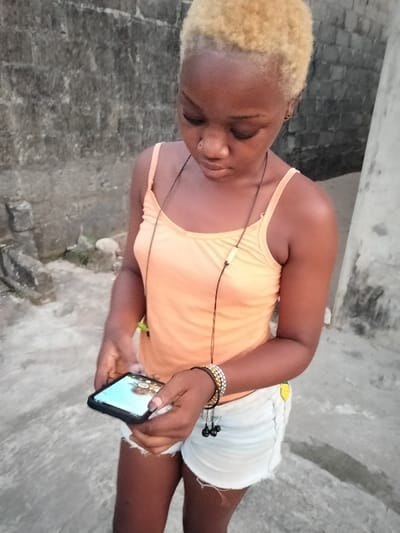 A young woman with bleached blonde hair stands outside, looking down at a phone she holds in her hands. She wears an orange tank top and denim shorts. A necklace hangs around her neck and beaded bracelets adorn her wrist, with earbuds hanging from them. The background shows a concrete wall and paved ground, suggesting an urban setting.