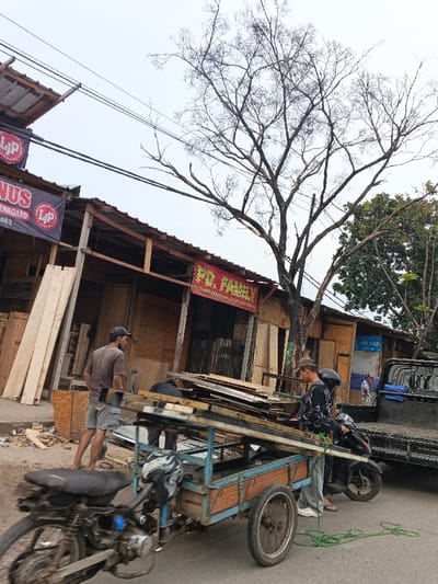 This image depicts a daytime street scene in Bandung City, Indonesia, focusing on commercial activity related to lumber or construction materials. The environment is characterized by an overcast sky, an asphalt road, and utility lines crisscrossing overhead.
On the left, a row of informal single-story shops or workshops lines the street. These structures feature facades made of wood planks and corrugated metal, supported by visible wooden frames. Several banners are affixed to the buildings; one on the far left partially displays "LJP PLYWOOD & PARTICLEBOARD," while a central red banner reads "PD FAUZIY." Further right, a blue sign indicates "PD. FIKAI." Piles of light-colored wood planks are stacked vertically outside the shops.
In the center of the frame, a prominent bare deciduous tree with numerous branches stands against the sky. To its right, a smaller tree with green foliage is visible.
Three adult males are central to the foreground activity. On the far left, a man in a dark t-shirt, light shorts, and a dark baseball cap stands barefoot, facing towards the right, appearing to observe the scene. In the mid-ground, a motorized three-wheeled cargo tricycle, equipped with a blue metal frame and wooden side panels, is laden with horizontal planks or metal frames. An individual in dark clothing is partially obscured behind these materials, seemingly engaged in handling them. To the right of the cargo tricycle, another man, wearing a patterned long-sleeve shirt, light pants, flip-flops, and a baseball cap, stands near a black scooter parked upright. He faces the cargo tricycle and the obscured individual, possibly supervising or assisting. A coiled green rope lies on the asphalt near the cargo tricycle. On the far right, a dark-colored pickup truck with a utility bed is parked, and a fourth individual in a patterned shirt is partially visible in the background near the truck.