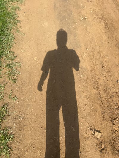 The image shows a long shadow of a person cast on a dirt path. The shadow is elongated due to the low angle of the sun, suggesting it is either early morning or late afternoon. The path is made of dry, sandy soil with some small stones and tire tracks visible, indicating it might be a road or a trail. In the lower part of the image, a strip of green grass is visible, suggesting the presence of vegetation alongside the path. The overall scene is outdoors, under a bright sky, typical of a sunny day. The focus is on the shadow, which takes up a significant portion of the frame, emphasizing the silhouette of a person. There is no visible text in the image. The context of Bauchi, Nigeria, implies a warm, likely dry climate during the time this photo was taken. The activity depicted is simply a person walking, as indicated by the shadow's form, though the person themselves is not visible, only their shadow. The mood is one of solitude and stillness, focusing on the play of light and shadow.