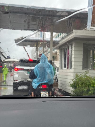 The image shows a street scene in Patong, Thailand, on a rainy day, as viewed from inside a vehicle. Raindrops are visible on the windshield, creating a blurred effect. In the foreground, the dashboard of the vehicle is visible. The middle ground features a motorcycle with two people on it, both wearing raincoats. The person in the back is wearing a light blue raincoat, and the person in front is wearing a dark helmet and appears to be driving. Behind the motorcycle is a white SUV with its taillights illuminated. To the left of the SUV, a person in a bright yellow raincoat is walking. In the background, a white building with a covered entrance and windows is visible, along with trees and other structures. The overall atmosphere is one of wet weather and traffic.