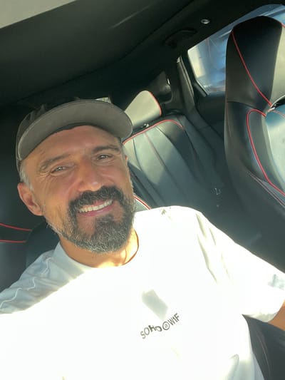 A close-up selfie shows a man in a car, wearing a gray baseball cap and a white t-shirt. The man has a beard and is smiling. The car's interior, with black leather seats and red stitching, is visible in the background. The scene is likely in Ko Samui, Thailand, given the bright sunlight, which suggests a tropical climate. The man appears to be relaxed and enjoying the drive.