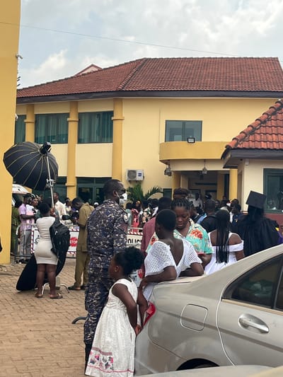 An outdoor scene in Madina, Ghana, depicts a densely packed crowd gathered in front of a multi-story building. The building, featuring a light-yellow facade with decorative columns and a brown tiled roof, displays multiple windows, some tinted, and an air conditioning unit on its exterior. A small balcony or portico extends from the building's entrance. In the foreground, a silver sedan is partially visible on the right. A young girl in a white patterned dress stands near the car, facing right. Adjacent to her, a woman in a white mesh-patterned top leans on the car. Centrally, a man in a blue camouflage uniform, wearing a white face mask and sunglasses, stands facing left. To the left, a woman in a light-colored fitted dress stands near professional photography equipment, which includes a large black umbrella diffuser on a stand. White crowd control barriers, partially obscuring text that reads "POLICE" and a red "STOP" sign, delineate areas within the gathering. A person wearing a black academic graduation gown and cap is visible on the far right. The background shows more people congregating, with hints of greenery near the building. The sky above is partly cloudy, and the ground is paved.