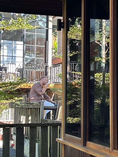An adult male is seated on a wooden balcony, appearing to smoke, with his right hand near his mouth. He is wearing a brown long-sleeved shirt and light-colored pants. The balcony overlooks a courtyard or outdoor area with lush green trees and foliage. In the background, a modern building with large windows and balconies is visible, along with other structures that suggest an urban or semi-urban setting. The scene is captured during daylight, with sunlight casting shadows and highlighting textures. The image is taken from a slightly elevated perspective, looking towards the man and the building. The specific location in Kediri, Indonesia, cannot be determined from the visual information alone.