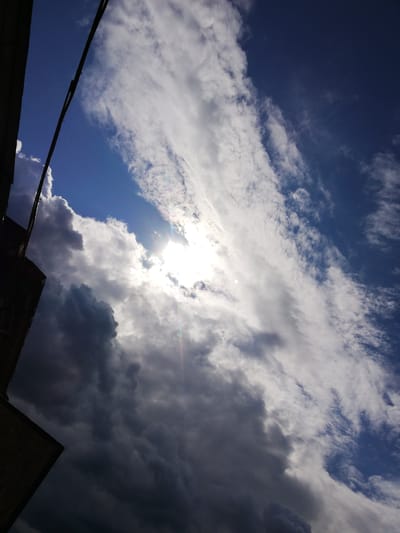 The image depicts a dramatic sky with a mix of dark, heavy clouds and bright, wispy clouds. The sun is partially obscured by the clouds, creating a bright glow and lens flare. In the foreground, the edge of a building or structure is visible on the left, silhouetted against the sky. A thin wire runs diagonally across the upper left portion of the image. The scene appears to be an outdoor view from a window or an elevated position. The general atmospheric conditions suggest a transition between stormy weather and clearer skies. The provided information states the location is Tosno, Russia, implying this is an urban or semi-urban landscape. There are no people or discernible actions visible in the image.