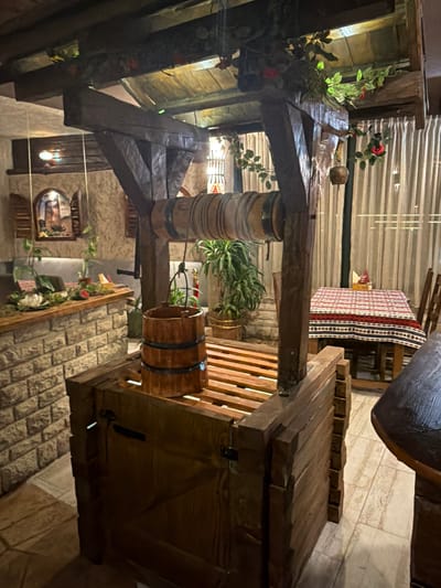 The image shows an indoor decorative well structure made of wood in a dimly lit restaurant or cafe. The well has a wooden bucket suspended from a rope and a crank mechanism. The structure is detailed with wooden planks and beams, and decorative vines with artificial red flowers are draped around it. In the background, there is a table set with a red and white patterned tablecloth, chairs, and a curtained window. To the left, a stone counter with plants and a painting on the wall is visible. The overall ambiance is rustic and possibly themed. This scene is located at a restaurant in Kumbor, Montenegro.