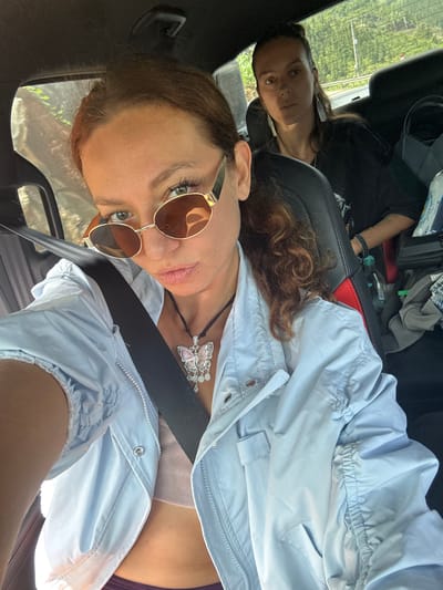 Two women are in a car. The woman in the foreground wears sunglasses, a light blue jacket, and a seatbelt. She is wearing a butterfly pendant. The other woman is in the background. The setting appears to be a roadside or highway based on the greenery visible through the car window. The specific location in Lũng Cú Commune, Vietnam, is difficult to determine without more contextual information.