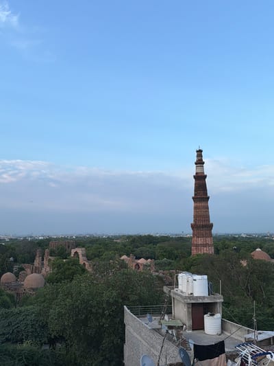 This is a high-angle, medium shot of the Qutub Minar in Delhi, India, seen from a rooftop at dusk. The Qutub Minar, a towering sandstone minaret, dominates the right side of the frame, its red and cream-colored bands visible against a light blue sky. In the background, lush green trees form a dense canopy over the surrounding landscape, interspersed with the ruins of older structures, including domed buildings and arches. A modern building with white water tanks and satellite dishes is in the foreground, occupying the lower right quadrant of the image. The overall scene depicts a blend of historical architecture and modern urban life, set against a backdrop of natural greenery.