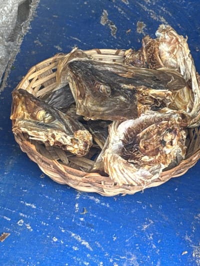 The image shows a woven basket filled with dried fish heads. The fish heads are brown and appear to be a whole fish, with visible eyes and scales. The basket is placed on a blue surface, which may be a table or a tarp. The background is a solid blue color, with some white speckles. The lighting is bright, suggesting it is daytime. The overall scene appears to be a market or a place where fish are sold or prepared. There are no people visible in the image. The location is Awka, Nigeria.