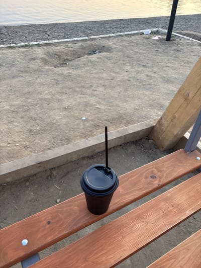 The image depicts a black disposable coffee cup with a straw placed on a wooden bench. The bench appears to be outdoors, situated on a sandy surface. In the background, a body of water with ripples is visible, bordered by a gravelly shore and a sandy area with some debris. A thin black pole, possibly a lamppost, is also present in the background. The lighting suggests it is either early morning or late afternoon. The scene appears to be a public area, potentially a park or a waterfront promenade in the city of Kemerovo, Russia. There are no people or any interactions visible in the image.