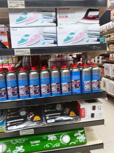 A store shelf displays an assortment of household items. The top shelf holds boxes of electric irons in pink and blue. Below, rows of blue gas canisters with red caps are visible. On the next shelf are packaged stove burners. The bottom shelf features a green Turbo stove with a floral pattern. Price tags are attached to some items. The store setting includes additional shelves in the background.