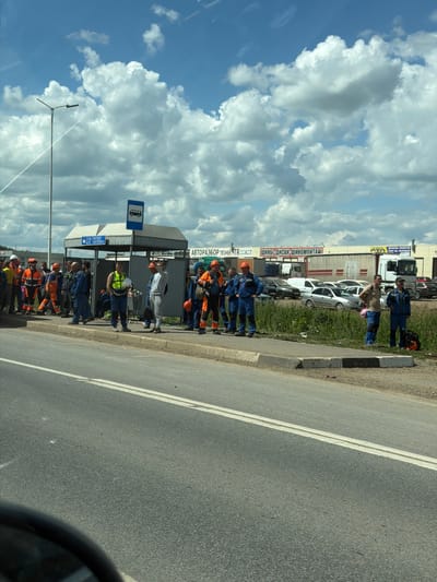 A group of male workers in various construction-style uniforms, some wearing high-visibility vests and hard hats, are gathered at a bus stop shelter. They are standing along a curb next to a road. Some are talking or looking towards the road, suggesting they are waiting for transportation. Behind the bus stop is a building with signage that reads "ШИНЫ-ДИСКИ-ШИНОМОНТАЖ" (Tires-Discs-Tire Fitting) and "АВТОРАЗБОР" (Auto Dismantling). There are vehicles, including trucks and cars, parked in front of the building. The scene is set under a bright, partly cloudy sky with a street lamp visible on the left. The ground is a mixture of asphalt and a grassy verge.