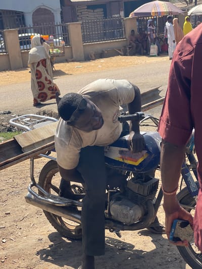 The image depicts a street scene in Zaria, Nigeria, under bright daylight. In the immediate foreground, a dark-skinned male, wearing a dirty white short-sleeved t-shirt, dark trousers, and sunglasses pushed onto his forehead, is bent over a blue motorcycle. His left hand is positioned on the motorcycle's fuel tank area, which exhibits wear and a partial white, yellow, and blue stripe design. The motorcycle has a visible engine block and exhaust pipe, and a metal rack on its rear carrying a rectangular light-colored object, possibly a piece of wood or metal. To the right, only the torso and arm of another individual, wearing a maroon long-sleeved garment and holding a blue smartphone with a patterned case, are visible. The ground is dusty and unpaved in the foreground.
In the midground, a paved road stretches across the frame. A woman dressed in a cream-colored hijab and a patterned long dress with orange, black, and white motifs walks away from the camera on the road. To the left of the road, a small vendor's cart with various bottled items is visible next to a black metal fence.
The background features a building with a light-colored facade, in front of which large sacks are stacked, likely containing grain or cement. Further behind, under a multi-colored striped umbrella, a group of individuals are seated and standing. Several other people, including women in hijabs and men in traditional attire, are visible walking or standing in the distance, suggesting a busy urban or market environment.