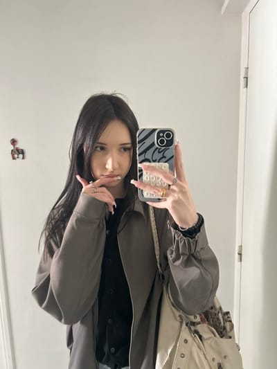 An individual, identified as a young female, is depicted from the mid-torso upward, reflected in a mirror within an indoor setting in Cachan, France. The subject has long, dark, straight hair with a center part and a light complexion. She wears a dark grey or taupe collared jacket, unzipped, over a black button-down garment. Her nails are long, manicured with white tips. She wears a silver-toned ring on her left ring finger, a gold-toned band on her right ring finger, and another silver-toned ring on her right pinky finger. A dark, coiled wristband is visible on her right wrist. She carries a light-colored shoulder bag or tote with visible metal embellishments and a patterned fabric protruding from its opening.
The subject is actively taking a mirror selfie, holding a smartphone in her right hand. The phone features a dark case with a grey and white abstract pattern and a multi-bubble pop-it style accessory attached to the back. Her left hand is raised, with fingers positioned near her lower lip. Her gaze is directed towards her left, away from the phone's camera lens.
The background consists of a plain, light-colored (white) wall. On the left side of the frame, a small, colorful decorative item resembling a house with a balloon or chimney is affixed to the wall. On the right, a portion of a white door frame and a silver-colored hinge are visible, indicating a closed door. The environment appears to be a private or semi-private interior space.