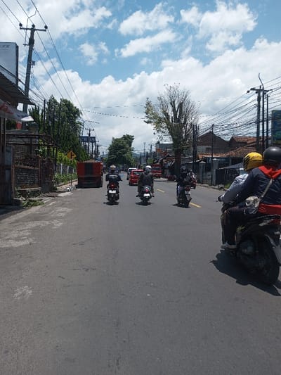 This daytime image captures a street scene in Tagtse Tashidingkha, Bhutan, under a blue sky with scattered white clouds. The asphalt road, which lacks visible lane markings, extends into the distance. In the foreground right, the rear of a dark Honda scooter is seen with two occupants; one wears a light shirt and dark pants, and a yellow 'MAXIM' helmet is visible near the driver. Four additional motorcycles, each with a single helmeted rider in dark clothing, proceed away from the viewer in the midground. Further ahead, a red truck with a dark bed is on the left, followed by two red cars. The left side of the street features a dark metal fence, buildings, dense green trees, multiple utility poles laden with numerous overhead wires, and a yellow right-turn arrow sign. The right side also presents buildings, utility poles, and a large tree with mostly bare branches. The scene is illuminated by strong sunlight, casting visible shadows.