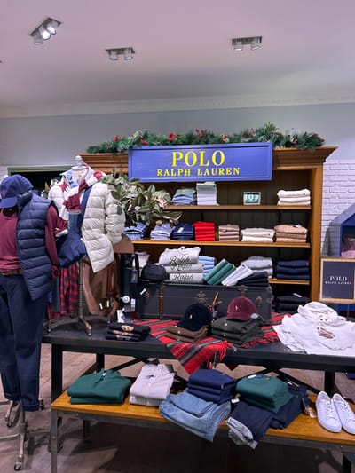 This image depicts an interior view of a Polo Ralph Lauren retail store located in Saint Julian's, Malta. The scene showcases a merchandise display, possibly seasonal, with garments arranged on various fixtures.
In the foreground, a lower metal-framed wooden table displays folded apparel including a green quarter-zip pullover, a striped long-sleeved shirt, several pairs of blue jeans, navy polo shirts, and a white long-sleeved shirt. A pair of white low-top sneakers is visible on the far right of this table.
Behind this, a larger, darker-toned display table features two headless mannequins. The mannequin on the left wears a dark blue baseball cap, a maroon long-sleeved polo shirt, a dark blue quilted vest, and dark blue trousers. The adjacent mannequin is dressed in a light beige quilted puffer jacket over a striped top, with a red plaid skirt or trousers, and a dark blue shoulder bag draped over its arm. A black belt is also displayed near this mannequin. The central part of this table is covered with a red and black plaid fabric, on which folded sweaters (including grey ones with "Ralph" text), dark blue and maroon baseball caps, and other folded apparel are arranged around a dark, trunk-like object with metallic accents.
In the background, a large wooden display hutch with multiple shelves is visible. A prominent blue sign with "POLO RALPH LAUREN" in yellow lettering is mounted on its upper section. The top of the hutch is decorated with a festive garland of green foliage and red berries. The shelves within the hutch are stocked with neatly folded sweaters and shirts in various colors, including blues, greens, reds, beiges, and greys. A small wooden sign with "POLO RALPH LAUREN" branding stands on the right side of the main display.
The store environment includes a light-colored ceiling with multiple recessed spotlights providing illumination. The walls feature a light grey or blue painted section on the left and a white brick-patterned section on the right. No individuals are visible within the frame.