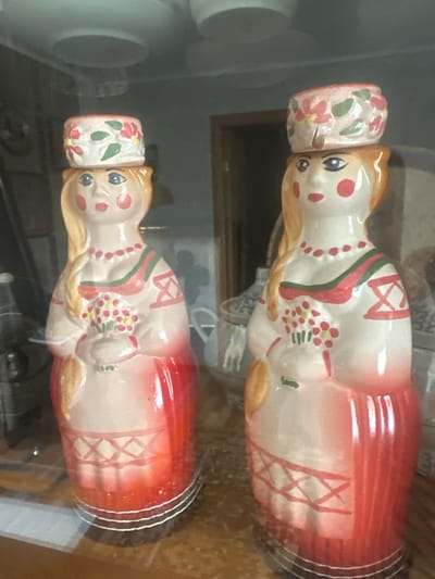 This media file shows two decorative ceramic figures, likely salt and pepper shakers, displayed on a shelf. The figures are identical and appear to be designed to resemble stylized human figures with red dresses and white bodices. They have painted facial features including blue eyes, red cheeks, and red lips. Each figure wears a decorative hat with a floral pattern. The dresses are predominantly red with white sections adorned with crisscross patterns. The figures are positioned side-by-side, with the image rotated. The background is blurred, showing a shelf and possibly other objects. The lighting suggests an indoor setting, possibly a home or shop. No text is visible in the image. The context provided indicates the location as Пярэжырскі сельскі Савет, Belarus.