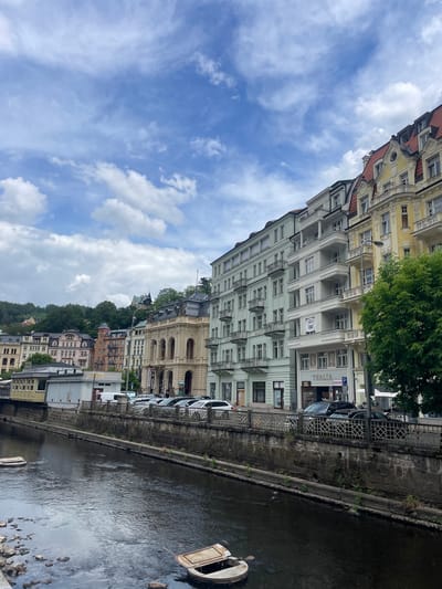 The image displays a daytime view of the Teplá River and its adjacent urban landscape in Karlovy Vary, Czechia. The sky, occupying the upper portion, is light blue with scattered white and grey clouds.

Along the right bank of the river, a row of multi-story buildings exhibits classic European spa town architecture. The leftmost visible building, partially obscured by a tree, is yellow with a red mansard roof and ornate white balconies. To its left, a light blue-green building with white balconies features a ground-floor storefront marked "THALIA" and "Style Shoes." Further left, another light green building displays "OPERA PALACE" on its facade. Beyond these, a grand, cream-colored building with large arched windows and a dark roof, possibly a theatre, has a "KONCERT" sign near its entrance. Several indistinct human figures are visible on the sidewalk in front of these buildings.

On the street running parallel to the river, numerous cars, including dark sedans and a white van, are parked. A metal fence with a lattice pattern separates the sidewalk from a stone riverbank wall, which has small plants growing from its crevices.

Further down the river, a diverse collection of buildings, varying in color from pink to light brown and white, line the riverbank and ascend a verdant, tree-covered hillside. One building clearly shows the sign "OLYMPIA." The distant hillside is densely forested, with hints of additional structures nestled within the trees.

The Teplá River flows through the lower-central part of the image. Its dark, reflective surface shows ripples and reflections of the sky and surrounding structures. On the immediate left bank, dark rocks are visible. Two white, boat-like or flat-topped structures are partially submerged or floating in the water, one in the lower-left corner and another in the lower-right. On the far left bank, a small yellowish building resembling a station or kiosk is visible next to a small white structure. The overall scene is illuminated by diffused daylight, indicative of a partly cloudy day.