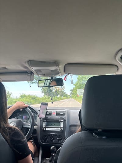 The image shows the interior of a car from a passenger's perspective, looking forward. A young woman is driving, her hands on the steering wheel, with her long dark hair falling over her shoulders. The car's dashboard is visible, featuring a radio and air conditioning controls. A smartphone is mounted on the dashboard, displaying a navigation app. A rearview mirror is positioned above the dashboard, reflecting the driver and the interior of the car.

Outside the car, trees and a road are visible through the windshield, suggesting a rural or suburban setting. The sky appears bright, indicating it's daytime. There are some decorative items hanging from the rearview mirror, including what looks like a string of charms and a red and white tassel.

Visible text includes "VW" on the steering wheel, and text on the radio display which is difficult to read clearly but includes frequency numbers like "87.5 MHz". A sticker on the windshield partially reads "IAVS ESSA".

The overall impression is that of a journey on a sunny day. The scene is mundane and focuses on the act of driving.