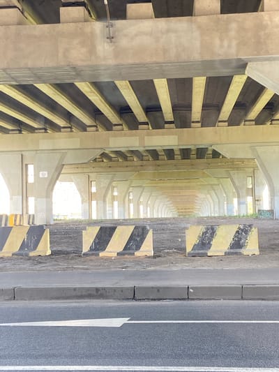 The image shows a view from under a bridge or overpass in the city of Shushary, Russia. The foreground features a road with a white directional arrow painted on it, indicating a lane. Adjacent to the road is a grey concrete curb. Behind the curb, there are several concrete barriers with black and yellow diagonal stripes, typical of traffic or construction zone safety. The ground behind the barriers is dirt and gravel.

The bridge structure itself is made of concrete. The underside of the bridge is composed of horizontal beams supported by vertical pillars. The pillars are spaced out, creating a tunnel-like effect that recedes into the distance. Some of the pillars have rectangular window-like openings. The overall impression is one of infrastructure, possibly in a suburban or industrial area. No people or animals are visible in the image. The lighting suggests it is daytime, with sunlight illuminating the scene from the side and the distance.