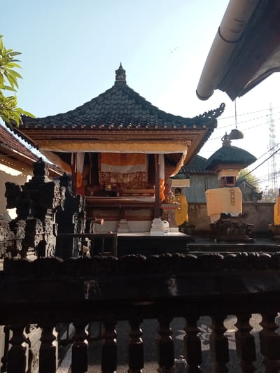 The image depicts a Balinese Hindu temple complex under a clear blue sky, likely in Blahbatuh, Indonesia, given the architectural style. The central focus is a tiered structure with a dark, multi-layered roof adorned with golden trim and decorative elements. Yellow and white fabric drapes are visible, adding to the sacred atmosphere. In the foreground, a dark, intricately carved stone fence runs horizontally across the frame, partially obscuring the view of the temple structures behind it. To the left, a lush green plant with leaves and small white flowers is visible, contrasting with the dark stone. To the right, another smaller shrine-like structure, also draped in yellow fabric, stands beside the main temple. The background shows other buildings and trees, and in the far distance, a telecommunications tower is faintly visible against the bright sky. The lighting suggests it is daytime, with sunlight casting shadows. There are no people or animals depicted in the scene. The overall impression is one of serene religious architecture amidst a tropical environment.