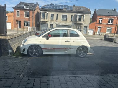 This image captures a bright, sunny daytime scene in what appears to be a residential area of Marche-en-Famenne, Belgium. The primary subject is a white Abarth car, parked facing left on a paved surface, likely a driveway or quiet street. The car features distinctive red side mirrors and a prominent red and white striped decal along its lower side, clearly displaying the text "ABARTH." It has dark, multi-spoke alloy wheels.
In the background, a row of European houses lines the street. The house immediately behind the car is constructed of red brick with dark window frames and a steeply pitched roof covered in numerous solar panels. To its right stands a lighter-colored, possibly stucco, building featuring several bay windows and also equipped with a significant array of solar panels on its roof. Further right, another red brick house with a grey roof is visible. The architecture, combining brick and stucco with various window styles and the prevalent use of solar panels, is characteristic of contemporary European residential areas.
The foreground consists of a mix of dark asphalt and grey cobblestone paving. The car is parked close to a low stone wall, behind which a small patch of grass is visible. The sky is clear blue, and strong sunlight casts distinct shadows, indicating a clear, bright day. No people are present, lending a quiet and still atmosphere to the scene.