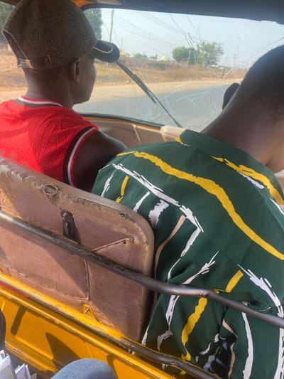 The image captures a scene inside a vehicle, likely a three-wheeled auto-rickshaw or similar utility vehicle, with two individuals present. The photo is taken from a slightly elevated, rear-facing perspective within the vehicle, looking forward.

In the foreground and center, a person is seated, presumably the driver, facing away from the camera. They are wearing a dark green shirt with prominent yellow, white, and light brown stripes. The fabric of the shirt appears somewhat creased. Their dark-skinned arm is visible on the left side of the frame. The seat they are on, partially visible below them, is upholstered in a worn, light brown or tan material, showing signs of aging and possibly damage, with some areas appearing cracked or discolored. A dark metal bar, possibly part of the vehicle's frame or handlebars, runs vertically in front of this seat.

Behind and to the left of the driver, the head and upper torso of a second person are visible. This individual has dark skin and is wearing a bright red sleeveless jersey, possibly a basketball jersey, with black and white trim around the neck and armholes. They are also wearing a dark grey or black cap. This person appears to be a passenger, seated behind the driver and looking forward.

The most striking detail is the vehicle's windshield, which is severely cracked. Numerous large cracks radiate across the glass, obstructing the view of the outside world. A windshield wiper is partially visible at the bottom edge of the cracked glass. Through the cracked windshield, a bright, sunny outdoor scene is visible, suggesting it is daytime with clear weather. A road or dirt path can be seen stretching into the distance, flanked by some sparse vegetation or trees. The exterior of the vehicle, particularly on the right side of the frame, shows parts of its bodywork in a bright yellow color. The general condition of the vehicle, particularly the cracked windshield and worn seats, suggests significant use or damage.