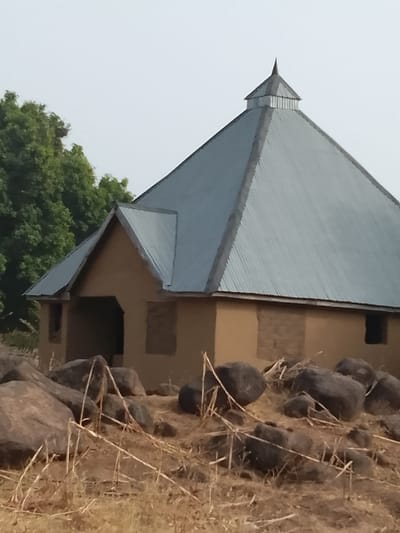 This image depicts a single-story building located in Jibam, Nigeria, under a light or hazy sky. The structure features light brown, possibly earthen or mud-plastered walls. Its roof comprises a prominent, steep, pyramidal section constructed from corrugated metal sheeting, topped with a small, pointed spire. An attached smaller section, also with a corrugated metal gable roof, contains an arched entrance opening. Several rectangular patches of unrendered or filled-in sections are visible on the building's walls. A small, dark, rectangular opening, possibly a window, is present on the right side of the main building. In the foreground, numerous large, dark grey-brown boulders are scattered across dry, sparse, light brown vegetation and stalks. Green foliage from trees is visible in the background to the left of the building. No individuals or active subjects are present.
