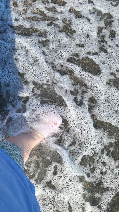 A low-angle shot shows a person's foot being washed by the foamy waves on a pebbly beach. The foot is adorned with pink nail polish and what appears to be a small, multi-colored charm on the big toe. The person is wearing a blue skirt. The scene is located on a beach in Khostinskiy, Russia.