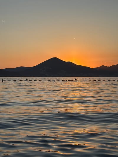 The image depicts a serene seascape at sunset, likely in the vicinity of Sveti Stefan, Montenegro. The horizon is dominated by the silhouette of rolling hills and mountains, with the sun partially obscured behind the central, largest peak, casting a warm, orange glow across the sky. The sky transitions from a pale blue at the top to a vibrant orange and yellow near the horizon.

In the foreground, the calm surface of the sea is visible, with gentle ripples reflecting the colors of the sunset, creating streaks of light and shadow on the water. A line of buoys or markers stretches horizontally across the middle ground, suggesting a designated swimming area or boundary. A few dark, indistinct figures, possibly people or objects, are visible near the buoys.

The overall atmosphere is tranquil and picturesque, capturing the beauty of a coastal sunset.