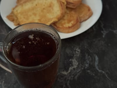 In Raivka, Ukraine, a close-up, eye-level shot features a transparent glass mug filled with a dark liquid, likely tea, with small bubbles on the surface. Behind the mug, on a white plate, are several pieces of golden-brown food, appearing to be French toast or fried bread. The food is arranged in a circular pattern. The background is a dark, textured surface, possibly a marble countertop, with subtle grey marbling. The lighting is dim, casting soft shadows and creating a slightly muted atmosphere.