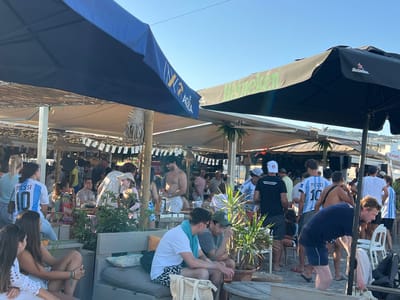 In San Roque, Spain, on a sunny day, a lively outdoor bar scene unfolds under a clear blue sky. Two large umbrellas, one navy blue and the other black with a white Heineken logo, provide shade. Underneath a straw-thatched roof structure, many people are gathered, some standing and others seated at tables. Several individuals are wearing Argentina national football team jerseys with "MESSI" and the number "10" on the back, suggesting a shared interest in football.
In the foreground, two young women are seated on a grey bench, with one in a white lace top and the other in a white tank top and denim shorts. To their right, two men are seated at a wooden table. One man in a white t-shirt and patterned shorts has a light blue towel draped over his shoulder, while the other wears a grey t-shirt and a dark green baseball cap. A beer glass sits on the table in front of them.
Further back, a shirtless man with white shorts is visible. Another man in a black t-shirt with white text is facing away from the camera. The background is filled with a crowd of people, some appearing to be in conversation or enjoying drinks. A tall plant with green leaves stands near the seated men, and another similar plant is positioned further back. The ground appears to be sand or a similar natural surface. The overall atmosphere suggests a casual, social gathering at a beachside establishment.