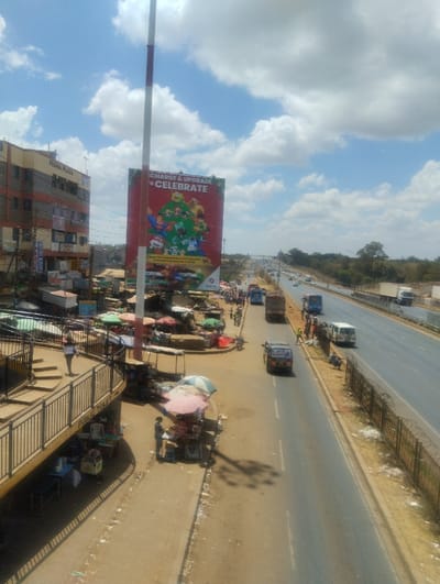 This elevated wide shot captures a bustling urban roadside environment in Witeithie ward, Kenya, under a bright, partly cloudy sky. In the immediate foreground, a metal-railed pedestrian overpass curves to the left, with a "PUBLIC TOILET" sign visible beneath. A lone individual stands on the overpass platform. Below the overpass, street vendors operate stalls, some shaded by light-colored umbrellas.
To the left, a multi-story building named "PEGRA PLAZA" rises above a dense market area populated by numerous vendor stalls, many covered with colorful tarps and umbrellas, where people are actively present. A tall red and white striped pole stands prominently in the center. Adjacent to this pole is a large vertical billboard, predominantly red, displaying "CHARGE & UPGRADE TO CELEBRATE" alongside various cartoon characters.
In the mid-ground, a two-lane asphalt service road runs parallel to the main highway. Vehicles, including a blue van, a brown truck, and a white van, are observed on this road, along with pedestrians. The background features a multi-lane highway carrying traffic in both directions, separated from the service road by a dirt median reinforced with a barbed-wire fence. Beyond the highway, the landscape transitions to a mix of trees and natural vegetation. Bright sunlight casts distinct shadows across the scene.